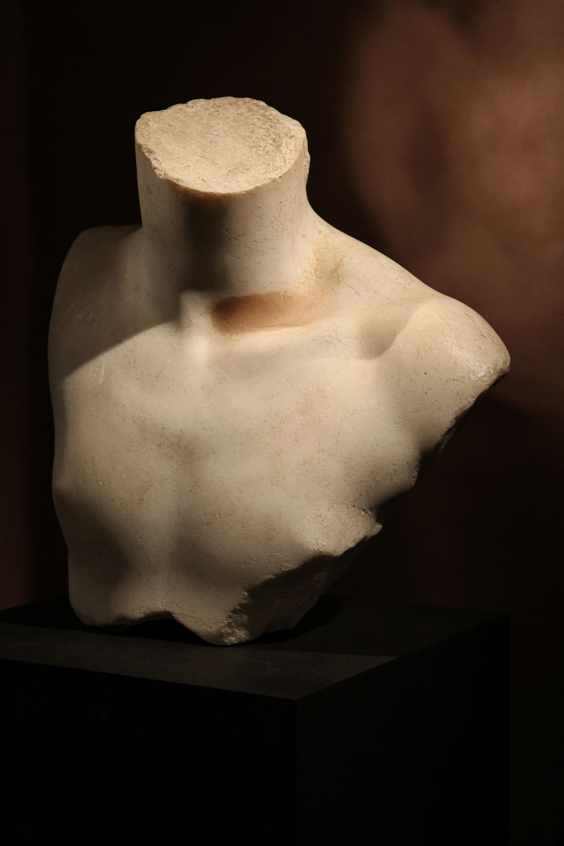 Pale marble fragment of a shoulder, collarbone and throat in raking museum light.