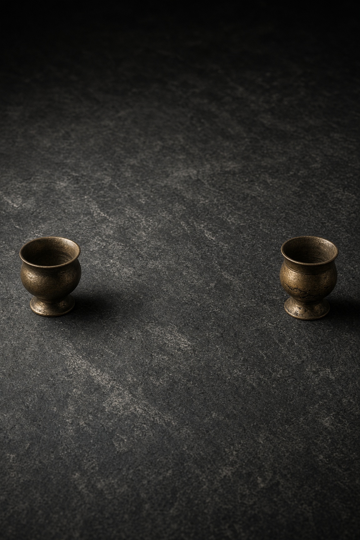 Two small bronze cups on dark stone with a wide empty interval between them.